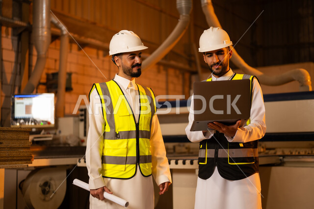 ورشات النجارة في السعودية، مهن ووظائف صناعية، معمل تصنيع الأثاث الخشبي، التحكم بالمعدات باستخدام التقنية والتكنولوجيا، المتابعة والإشراف على آلات المعمل، مهندس عربي خليجي سعودي يرتدي سترة وخوذة الحماية يحمل بيده الحاسوب المحمول يقف برفقة زميله