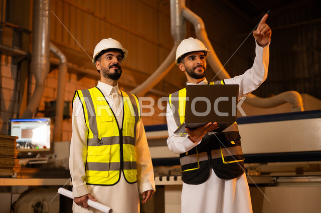ورشات النجارة في السعودية، مهن ووظائف صناعية، معمل تصنيع الأثاث الخشبي، التحكم بالمعدات باستخدام التقنية والتكنولوجيا، المتابعة والإشراف على آلات المعمل، مهندس عربي خليجي سعودي يرتدي سترة وخوذة الحماية يحمل بيده الحاسوب المحمول يقف برفقة زميله