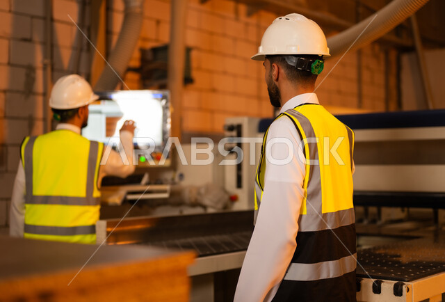 ورشات النجارة في السعودية، مهن ووظائف صناعية، معمل تصنيع الأثاث المنزلي، اجهزة ومعدات ضخمة، صورة من الجانب لمهندس عربي خليجي سعودي يرتدي الخوذة وسترة الحماية يقوم بالاشراف على سير خطة العمل