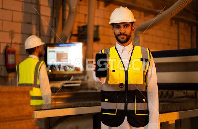 عرض شاشة سوداء فارغة، ورشات النجارة في السعودية، المتابعة والإشراف على آلات المعمل، التحكم بالمعدات باستخدام التقنية والتكنولوجيا، معمل تصنيع الأثاث الخشبي، مهن ووظائف صناعية، مهندس عربي خليجي سعودي يرتدي سترة وخوذة الحماية يحمل بيده الهاتف المحمول