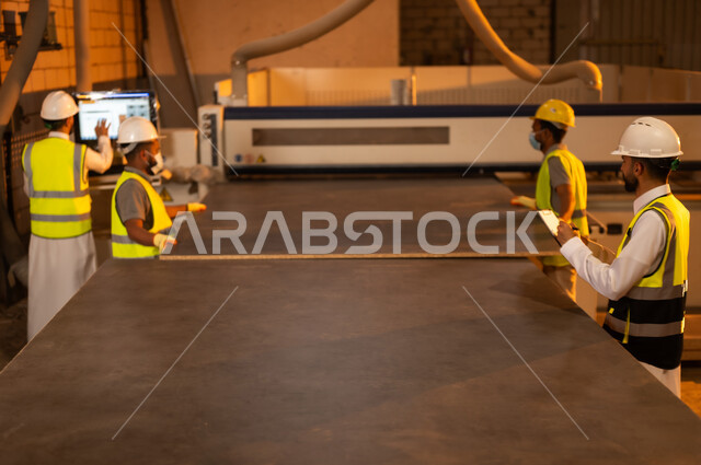 Industrial jobs and professions, laser printing on wood pieces, computer woodworking machine, laser engraving machine, taking notes on clipboard, group of Saudi Arabian Gulf craftsmen wearing protective jacket and helmet working in a carpentry workshop in Saudi Arabia