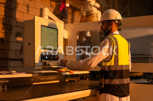 العمل في القطاع الصناعي, صورة مقربة من الجانب لمهندس عربي خليجي سعودي يرتدي الخوذة وسترة الحماية يعمل على آلة الطباعة الليزرية, اختيار الرسومات والنقوش عبر الحاسب الآلي, التطور والتقدم التقني في صناعة الخشب، معمل تصنيع الأثاث المنزلي