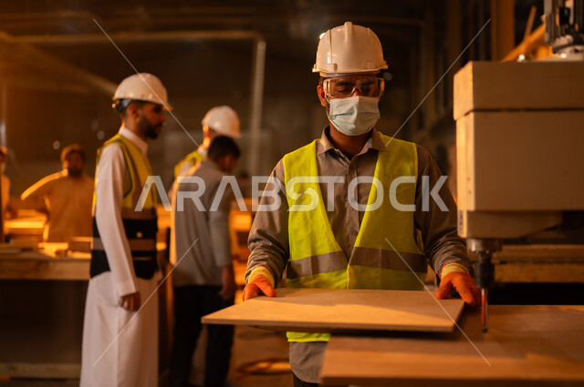 Working in the industrial sector, development and technical progress in the wood industry, two Saudi Arabian Gulf engineers wearing helmets and protective vests supervising workers, home furniture manufacturing plant, a worker working on a laser wood cutting machine