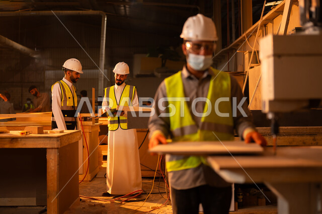 العمل في القطاع الصناعي، التطور والتقدم التقني في صناعة الخشب، مهندسين عربيين خليجيين سعوديين يرتديان الخوذة وسترة الحماية يقومان بالاشراف على العمال، معمل تصنيع الأثاث المنزلي، عامل يقوم بالعمل على جهاز قص الخشب بالليزر