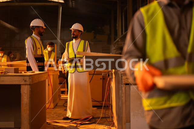 الاشراف على العمال في الورشة، متابعة سيرالعمل، ورشات النجارة في السعودية، معمل تصنيع الأثاث الخشبي، مهن ووظائف صناعية، مهندس عربي خليجي سعودي يرتدي سترة وخوذة الحماية يتحدث مع عامل