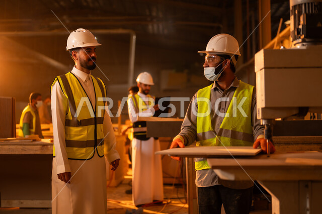 ورشات النجارة في السعودية، الاشراف على العمال في الورشة، متابعة سيرالعمل، معمل تصنيع الأثاث الخشبي، مهن ووظائف صناعية، مهندس عربي خليجي سعودي يرتدي سترة وخوذة الحماية يتحدث مع عامل