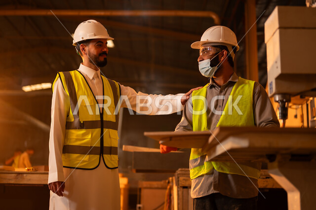 متابعة سيرالعمل، الاشراف على العمال في الورشة، ورشات النجارة في السعودية، معمل تصنيع الأثاث الخشبي، مهن ووظائف صناعية، مهندس عربي خليجي سعودي يرتدي سترة وخوذة الحماية يضع يده على كتف العامل