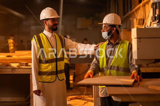 Follow up the workflow, supervise the workers in the workshop, carpentry workshops in Saudi Arabia, wooden furniture manufacturing plant, industrial professions and jobs, a Saudi Arabian Gulf engineer wearing a protective jacket and helmet puts his hand on the worker's shoulder