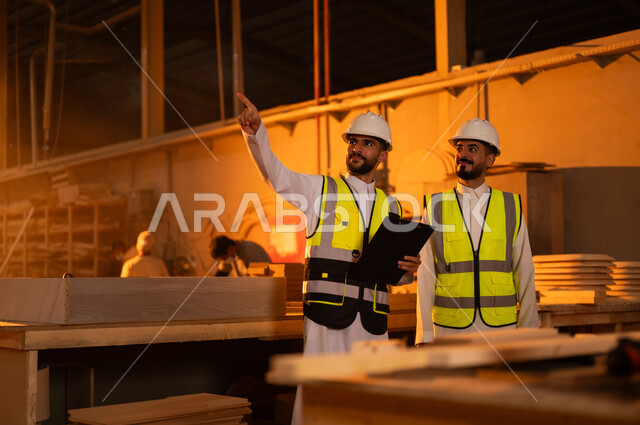 تسجيل وتدوين الملاحظات، المتابعة والإشراف على آلات المعمل، ورشات النجارة في السعودية، معمل تصنيع الأثاث الخشبي، مهن ووظائف صناعية، مهندس عربي خليجي سعودي يرتدي سترة وخوذة الحماية يحمل بيده حافظة ورقية يقف برفقة زميله