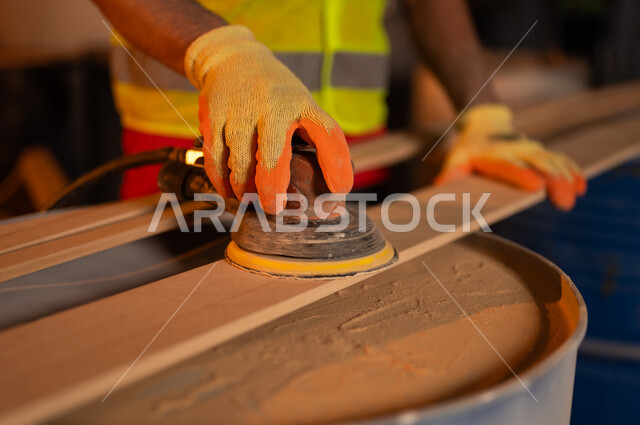 استخدام ادوات و معدات النجارة، صورة مقربة ليد حرفي عربي خليجي سعودي  يرتدي سترة الحماية وقفازات يقوم ببرد و تسوية قطعة الخشب في منجرة، ورشة ومصنع عمل، حرف ومهن عربية سعودية تقليدية قديمة