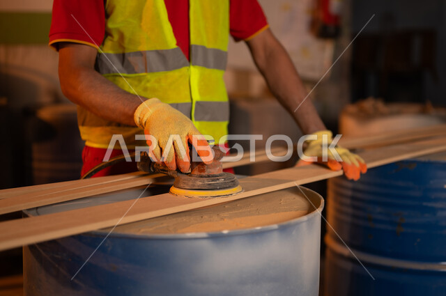 ورشة ومصنع عمل، استخدام ادوات و معدات النجارة، صورة مقربة لحرفي عربي خليجي سعودي  يرتدي سترة الحماية وقفازات يقوم ببرد و تسوية قطعة الخشب في منجرة، حرف ومهن عربية سعودية تقليدية قديمة