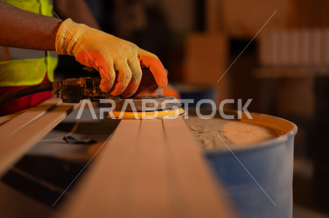 استخدام ادوات و معدات النجارة، صورة مقربة ليد حرفي عربي خليجي سعودي  يرتدي سترة الحماية وقفازات يقوم ببرد و تسوية قطعة الخشب في منجرة، ورشة ومصنع عمل، حرف ومهن عربية سعودية تقليدية قديمة