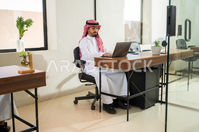 إرسال بريد الكتروني عبر الحاسوب, دمج التقنيات الحديثة و المتطورة مع العمل, مهن و أعمال مكتبية إدارية, التواصل مع العملاء عن بعد, شاب عربي خليجي سعودي يرتدي الثوب التقليدي و الشماغ يجلس داخل المكتب و يقوم بالكتابة على لوحة المفاتيح