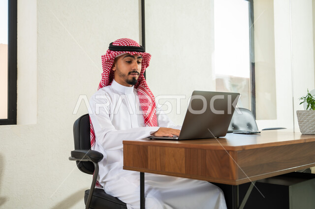دمج التقنيات الحديثة و المتطورة مع العمل, مهن و أعمال مكتبية إدارية, التواصل مع العملاء عن بعد, شاب عربي خليجي سعودي يرتدي الثوب التقليدي و الشماغ يجلس داخل المكتب و يقوم بالكتابة على لوحة المفاتيح, إرسال بريد الكتروني عبر الحاسوب