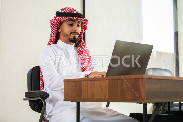 مهن و أعمال مكتبية إدارية, التواصل مع العملاء عن بعد, استخدام التقنيات الحديثة و المتطورة في العمل, شاب عربي خليجي سعودي يرتدي الثوب التقليدي و الشماغ يجلس داخل المكتب و يقوم بالكتابة على لوحة المفاتيح, إرسال بريد الكتروني عبر الحاسوب
