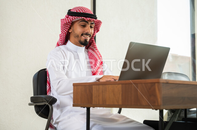 التواصل مع العملاء عن بعد, مهن و أعمال مكتبية إدارية, استخدام التقنيات الحديثة و المتطورة في العمل, شاب عربي خليجي سعودي يرتدي الثوب التقليدي و الشماغ يجلس داخل المكتب و يقوم بالكتابة على لوحة المفاتيح, إرسال بريد الكتروني عبر الحاسوب