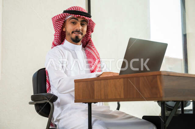 النظر إلى الكاميرا بإيماءات السعادة و السرور, إرسال بريد الكتروني عبر الحاسوب, استخدام التقنيات الحديثة و المتطورة في العمل, شاب عربي خليجي سعودي يرتدي الثوب التقليدي و الشماغ يجلس داخل المكتب و يقوم بالكتابة على لوحة المفاتيح, التواصل مع العملاء عن بعد