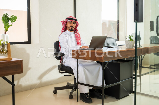 النظر إلى الكاميرا بإيماءات السعادة و السرور, إرسال بريد الكتروني عبر الحاسوب, استخدام التقنيات الحديثة و المتطورة في العمل, شاب عربي خليجي سعودي يرتدي الثوب التقليدي و الشماغ يجلس داخل المكتب و يقوم بالكتابة على لوحة المفاتيح, التواصل مع العملاء عن بعد