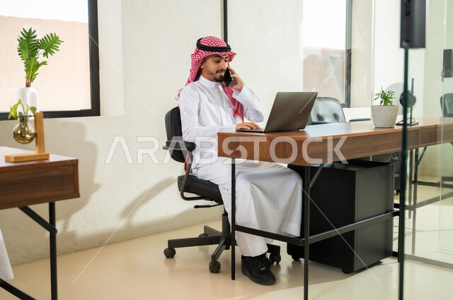 مهن و أعمال إدارية مكتبية, إجراء اتصال هاتفي عبر الجوال, استخدام التقنيات الحديثة و المتطورة في العمل, شاب عربي خليجي سعودي يرتدي الثوب التقليدي و الشماغ يجلس في المكتب و يمسك الهاتف المحمول بيده, التواصل مع العملاء عن بعد
