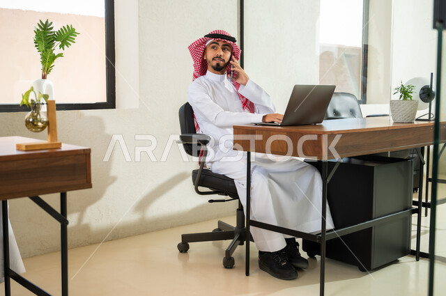 استخدام التقنيات الحديثة و المتطورة في العمل, مهن و أعمال إدارية مكتبية, إجراء اتصال هاتفي عبر الجوال, شاب عربي خليجي سعودي يرتدي الثوب التقليدي و الشماغ يجلس في المكتب و يمسك الهاتف المحمول بيده, التواصل مع العملاء عن بعد