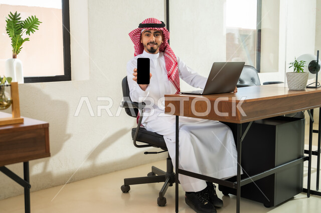Professions and office administrative work, using modern and advanced technologies at work, a Saudi Gulf Arab young man wearing the traditional dress and shemagh sitting inside the office and holding the mobile phone in his hand, showing a blank black screen on the mobile phone