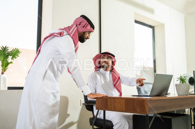تبادل الخبرات و المهارات, مفهوم الشراكة والتعاون, موظفان عربيان خليجيان سعوديان يعملان داخل مكتب, استخدام الأجهزة والتقنيات الحديثة في الأعمال المكتبية، إنجاز وتحقيق مهام العمل, شاب يشير بإصبع السبابة إلى شاشة الحاسوب
