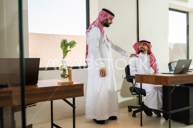 مفهوم التعاون بين الزملاء, أعمال إدارية و مكتبية, مهن و وظائف في السعودية, شاب عربي خليجي سعودي يرتدي الثوب التقليدي و الشماغ يضع يده على كتف صديقه بتعابير الدعم و المساندة, دمج التقنيات الحديثة بالعمل