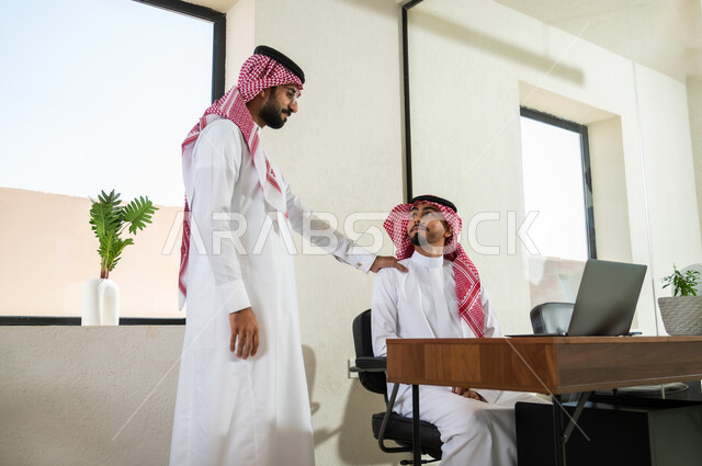 مفهوم التعاون بين الزملاء, أعمال إدارية و مكتبية, مهن و وظائف في السعودية, شاب عربي خليجي سعودي يرتدي الثوب التقليدي و الشماغ يضع يده على كتف صديقه بتعابير الدعم و المساندة, دمج التقنيات الحديثة بالعمل