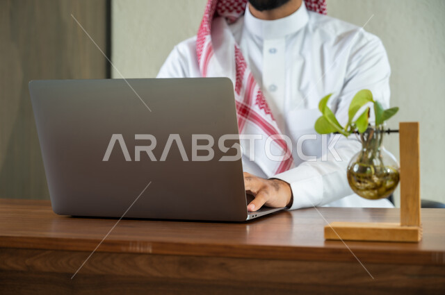 مهن و أعمال إدارية مكتبية, إرسال بريد الكتروني عبر الحاسوب, استخدام التقنيات الحديثة و المتطورة في العمل, صورة مقربة لشاب عربي خليجي سعودي يرتدي الثوب التقليدي و الشماغ يجلس داخل المكتب و يقوم بالكتابة على لوحة المفاتيح, التواصل مع العملاء عن بعد