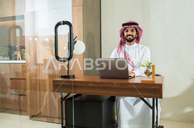 النظر إلى الكاميرا بإيماءات السعادة و السرور, إرسال بريد الكتروني عبر الحاسوب, دمج التقنيات الحديثة و المتطورة مع الأعمال المكتبية, شاب عربي خليجي سعودي يرتدي الثوب التقليدي و الشماغ يجلس داخل المكتب و يقوم بالكتابة على لوحة المفاتيح, التواصل مع العملاء عن بعد