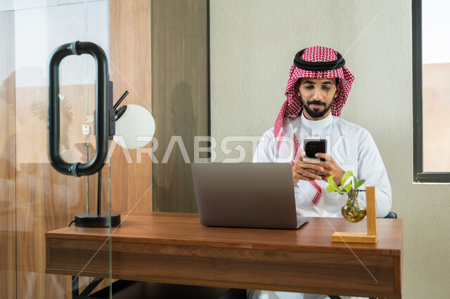 التواصل مع العملاء عن بعد, استخدام التقنيات الحديثة و المتطورة في العمل, مهن و وظائف في السعودية, شاب عربي خليجي سعودي يرتدي الثوب التقليدي يجلس داخل المكتب و يمسك الهاتف المحمول بيده, أعمال إدارية مكتبية