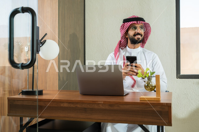 Communicating with clients remotely, using modern and advanced technologies at work, professions and jobs in Saudi Arabia, a Saudi Gulf Arab young man wearing a traditional dress sitting inside the office and holding a mobile phone in his hand, office administrative work