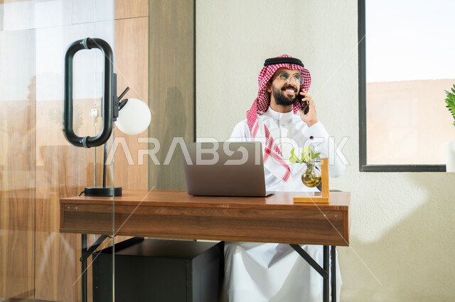 إجراء اتصال هاتفي عبر الجوال, استخدام التقنيات الحديثة و المتطورة في العمل, مهن و أعمال مكتبية في السعودية, شاب عربي خليجي سعودي يرتدي الثوب التقليدي و الشماغ  يجلس في المكتب و يمسك الهاتف المحمول بيده, التواصل مع العملاء عن بعد