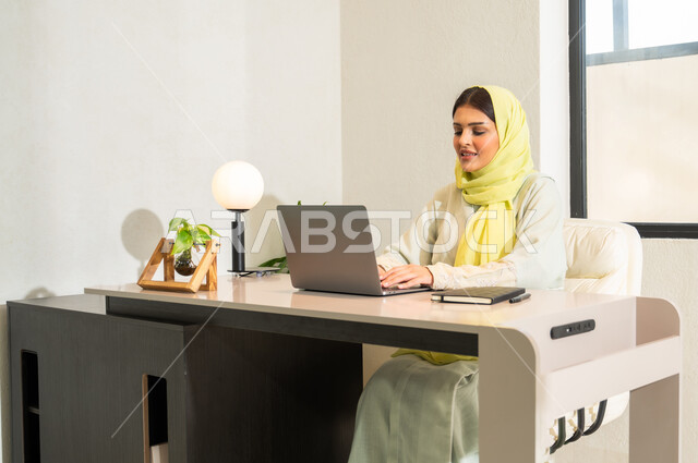 Sending e-mail via computer, using modern and advanced technologies at work, office professions and work in Saudi Arabia, a veiled Saudi Gulf Arab woman wearing a colorful abaya sitting in the office and typing on the keyboard, communicating with clients remotely.