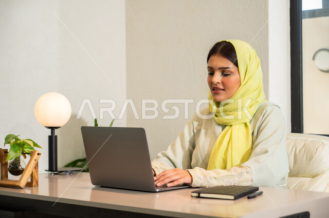 التواصل مع العملاء عن بعد, إرسال بريد الكتروني عبر الحاسوب, استخدام التقنيات الحديثة و المتطورة في العمل, مهن و أعمال مكتبية في السعودية, امرأة عربية خليجية سعودية محجبة ترتدي العباءة الملونة تجلس في المكتب و تقوم بالكتابة على لوحة المفاتيح
