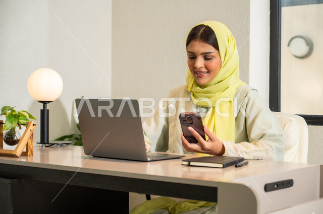 Communicating with clients remotely, using modern and advanced technologies at work, office professions and work in Saudi Arabia, a veiled Saudi Gulf Arab woman wearing a colorful abaya sitting in the office and holding a mobile phone in her hand, sending an email via the computer
