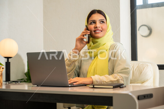 Making a phone call via mobile phone, using modern and advanced technologies at work, professions and office work in Saudi Arabia, a close-up photo of a veiled Saudi Arabian Gulf woman wearing a colorful abaya, sitting in the office and holding the mobile phone in her hand, communicating with clients remotely.