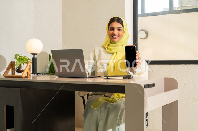استخدام التقنيات الحديثة و المتطورة في العمل, مهن و أعمال مكتبية في السعودية, امرأة عربية خليجية سعودية محجبة ترتدي العباءة الملونة تجلس في المكتب و تمسك الهاتف المحمول بيدها, عرض شاشة سوداء فارغة عبر الجوال