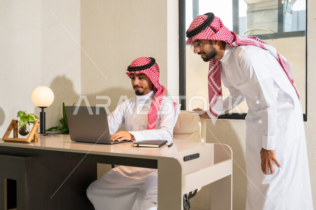 Integrating modern techniques and technology with office work, exchanging experiences and skills, two young Saudi Gulf Arabs wearing the traditional thobe and shemagh discuss work tasks.