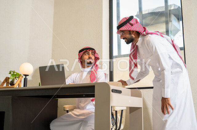 تبادل الخبرات و المهارات، دمج التقنيات الحديثة و التكنولوجيا بالأعمال المكتبية، شابان عربيان خليجيان سعوديان يرتديان الثوب التقليدي و الشماغ يقومان بمناقشة مهام العمل