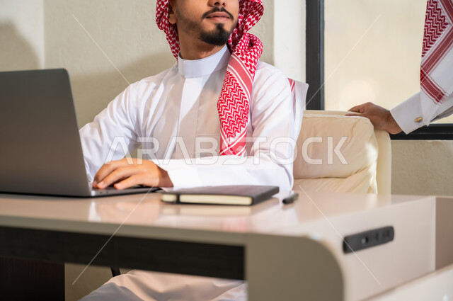 Exchanging experiences and skills, integrating modern technologies and technology into office work, a close-up photo of two young Saudi Gulf Arabs wearing the traditional thobe and shemagh discussing work tasks.