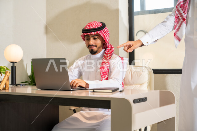 مفهوم التعاون والتوجيه, تبادل الخبرات و المهارات، دمج التقنيات الحديثة و التكنولوجيا بالأعمال المكتبية، شابان عربيان خليجيان سعوديان يرتديان الثوب التقليدي و الشماغ يقومان بمناقشة مهام العمل