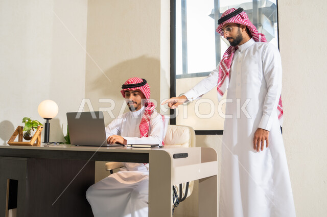 مفهوم التعاون والتوجيه, تبادل الخبرات و المهارات، دمج التقنيات الحديثة و التكنولوجيا بالأعمال المكتبية، شابان عربيان خليجيان سعوديان يرتديان الثوب التقليدي و الشماغ يقومان بمناقشة مهام العمل
