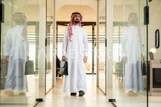 بيئة عمل مريحة في الشركات السعودية، النظر إلى شيء ما بإيماءات الفخر، مهن ووظائف مكتبية، رجل اعمال عربي خليجي سعودي يرتدي الزي التقليدي السعودي يمسك بيده حقيبة يمشي في مقر الشركة، الاستعداد لبدء يوم عمل جديد