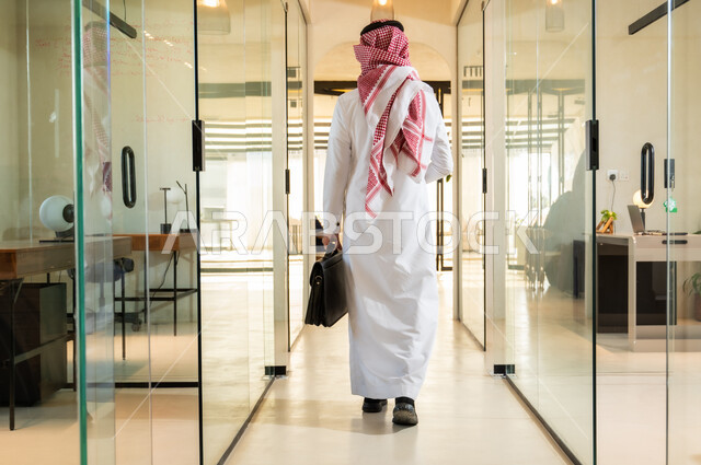 مهن ووظائف مكتبية في المملكة العربية السعودية، بيئة عمل مريحة في الشركات السعودية، صورة من الخلف لرجل اعمال عربي خليجي سعودي يرتدي الزي التقليدي السعودي يمسك بيده حقيبة يمشي في مقر الشركة، الاستعداد لبدء يوم عمل جديد