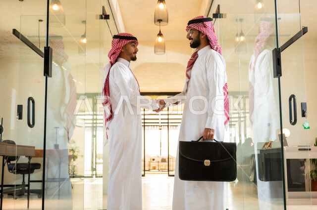 Exchange of experiences between the team to achieve goals and develop plans, office jobs and professions in the Kingdom of Saudi Arabia, comfortable work environment in Saudi companies, two Saudi Gulf Arab businessmen wearing traditional Saudi dress holding a bag in their hands standing at the company headquarters,