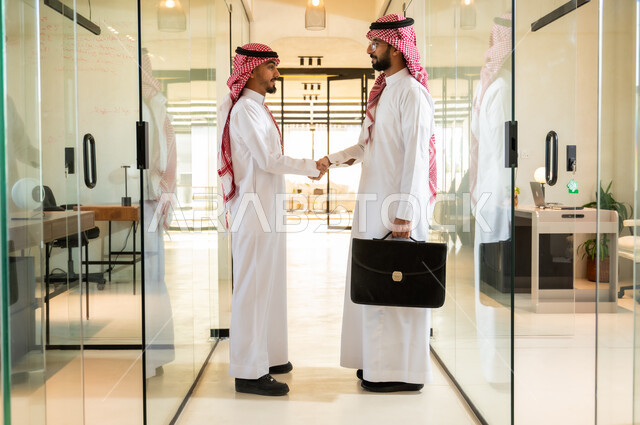 Office jobs and professions in the Kingdom, greetings, peace and agreement on successful business deals, a high-end work environment in Saudi companies, two Saudi Gulf Arab businessmen wearing traditional thobe and shemagh standing in the company headquarters shaking hands with expressions of joy and happiness