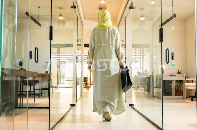 الاهتمام بالمظهر الخارجي، مهن ووظائف مكتبية في المملكة العربية السعودية، بيئة عمل مريحة في الشركات السعودية، صورة من الخلف لسيدة اعمال عربية خليجية سعودية ترتدي العباية والحجاب تمسك بيدها حقيبة تمشي في مقر الشركة، الاستعداد لبدء يوم عمل جديد