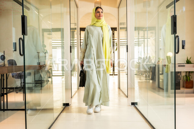 الاستعداد لبدء يوم عمل جديد، الاهتمام بالمظهر الخارجي، مهن ووظائف مكتبية في المملكة العربية السعودية، بيئة عمل مريحة في الشركات السعودية، سيدة اعمال عربية خليجية سعودية ترتدي العباية والحجاب تمسك بيدها حقيبة تمشي في مقر الشركة