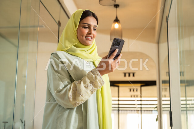 مهن ووظائف نسائية مكتبية، النظر الى الكاميرا بإيماءات البهجة والسرور، استخدام الأجهزة والتقنيات الحديثة في إنجاز المهام, استعمال تطبيقات وخدمات الهاتف المحمول, شابة عربية خليجية سعودية ترتدي العباية والحجاب تقف داخل مقر الشركة تمسك الجوال وتقوم بقراءة رسالة ما